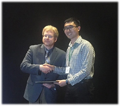Two men shaking hands in front of a black background, holding an award